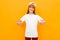 Red-haired charming girl in a white T-shirt on a background of an orange wall, points her fingers to herself, mock-up