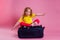Red-haired female child striped yellow t-shirt sitting on a large travel suitcase dream flies rest on the sea shore pink