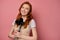 A red-haired girl in a apron and a T-shirt stands on a pink background, clutching kitchen appliances to chest and