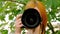 Red-haired girl holding a camera in the summer against a background of green leaves. A large lens is close-up aimed at the camera
