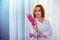 Red-haired girl manicure master stands against the backdrop of a gray-blue room wall and holds a manicure tool