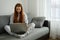 Red-haired girl in pajamas and glasses sits on a sofa and types on a laptop