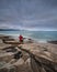 Red-Haired Girl-Photographer In Red Waistcoat On The Stony Shore Of Turquoise Sea Takes Pictures Of The Dawn. Girl And The Sea. Fe