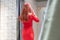 Red-haired girl in a red dress looks in the mirror. female in a fitting room for clothes. view from the back, slim figure of a