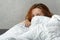 Red-haired girl shyly peeks out from under the covers. Portrait of a beautiful young women