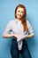 Red-haired girl in a striped shirt stands on a blue background and smiles, knotting the edges of the shirt