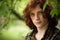 Red-haired man posing in sunny day outdoors. Young foxy guy in a plaid shirt looks at camera standing against on green