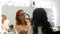 Red-haired stylist make-up artist doing stylish evening make-up of model with long black hair in beauty salon