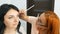 Red-haired stylist make-up artist doing stylish evening make-up of model with long black hair in beauty salon