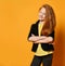 Red-haired teenage girl in black jacket, pants, yellow t-shirt. She smiling, hands folded, posing on orange background. Close up