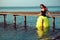 Red haired woman in black corset and long tail green veiling skirt standing in sea water and leaning on wooden pier.