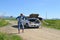 Red-haired woman pointing finger to the broken car with opened hood and brunette woman on a country road and barrier with stop sig