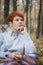 Red-haired woman sitting in a folding deck chair sheltered by a bright woolen plaid on a blurred background of an autumn forest,