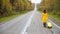 A red-haired woman in a yellow coat walks with a yellow suitcase along the highway in autumn.