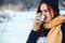 Red-haired young woman in scarf drinking tea with thermos in winter forest.