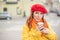 The red-haired young woman in a yellow coat and red beret is drinking coffee on the street to keep warm