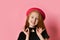 Red-headed teen female in black dress and red hat. She smiling, holding on to choker, posing on pink studio background. Close up