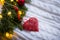 Red heart with ornament near fir wreath decorated with Christmas balls and coiled with glowing garland with warm light on white kn