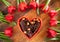 Red heart shaped bowl with chocolate and red tulips lying in around, on diagonal wooden background.