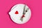 Red heart on a white plate  a fork  a knife  a red ribbon on a pink background  top view  copy space. Wedding  Valentine`s day