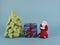 A red knitted angel stands next to a low Christmas tree. Between them lies a New Year\\\'s gift tied with a red ribbon