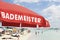 Red lifeguard umbrella on a beach against blue sky. Some bathers ont he background. The text on the umbrella Bademeister