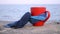 Red mug with coffee tied with blue knitted scarf stands on sandy beach