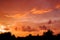 Red and orange cloudy sky before sunrise against a black silhos of rustic roofs and trees.