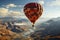 Red orange hot air balloon flies against a partly cloudy sky above a river through a beautiful valley with a backdrop of mountains