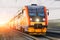 Red orange passenger train with headlights on rushing through the railway in the evening.