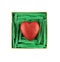 Red painted polyfoam heart enclosed with green paper raffia strips in beige box isolated on white background