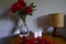 Red peonies in glass vase, straw lamp, white candles on wooden commode across white wall. Interior design. Home decor. lights and