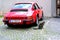 Red Porsche car parked near house in historic district old european city, young cat whiskas color sits on square, concept survival