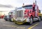 Red powerful big rig semi truck with oversize load sign on the grill guard bumper standing with loaded semi trailer at row on the