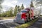 Red powerful bonnet classic American big rig semi truck with chrome parts and dry van semi trailer parked on the street with trees