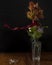 A red ribbon is moved by the wind around a glass vase containing flowers and leaves on a wooden table with vintage scissors