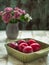 Red ripe plums lie in a deep square ceramic olive plate on a light wooden table. In the background, a small bouquet of