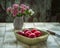 Red ripe plums lie in a deep square ceramic olive plate on a light wooden table. In the background, a small bouquet of