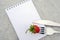 Red ripe strawberries berry on fork, knife and white blank notepad on gray stone background table. Top view, flat lay, Mockup.