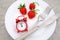 Red ripe strawberries berry on white plate, cutlery and red alarm clock on gray stone background table, closeup. Top view, flat