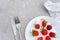 Red ripe strawberries berry on white plate, cutlery and red alarm clock on gray stone background table. Top view, flat lay, copy
