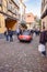 Red sports car drives through town with tourists onlooking.