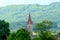 The red steeple of a church is seen against forest-covered hills.