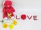 Red teddy bear sit hold the red heart with LOVE lettering made of red seeds and red box of the ring with flowers on white backgrou