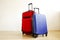 Red textile suitcase & blue hard shell luggage with extended telescopic handle up on wooden floor, white wall background. Couples