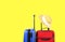 Red textile suitcase & blue hard shell luggage with straw hat hanging on extended telescopic handle, bright yellow backgr