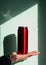 Red thermos can bottle in male hands against wall with harsh shadow. Hot tea coffee drinking in cold autumn winter weather