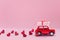Red toy car delivering carrying on roof gift box with small red hearts on pink background. Valentine`s day concept