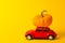 Red toy car and orange pumpkin on the roof on the yellow background. Red vintage toy car with small Halloween pumpkins on the roof