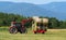 Red tractor loads round hay bales on trailer in a early summer day